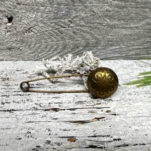 Könnte beinhalten: Vintage Sicherheitsnadel aus Messing mit einem verzierten, runden Knopf. Der Knopf weist ein detailliertes florales Design auf. Die Nadel befindet sich auf einer verwitterten, weiß lackierten Holzoberfläche mit einem Zweig aus getrocknetem weißem Laub.