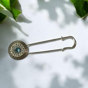 Könnte beinhalten: Silberfarbene Sicherheitsnadel mit einem verzierten, kreisförmigen Kopf. Der Kopf weist ein detailliertes, geprägtes Design um einen zentralen, hellblauen Edelstein auf. Die Nadel liegt vor einem weißen Hintergrund mit Schatten und grünen Blättern.