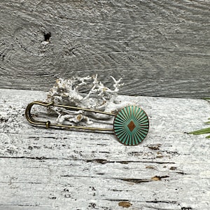 Könnte beinhalten: Eine Sicherheitsnadel im Vintage-Stil mit einem runden, türkisfarbenen Dekorationselement. Die Nadel hat eine Bronzefinish und ein Sonnenstrahlendesign, das von einer zentralen Diamantenform ausgeht. Vor einem verwitterten, weißen Holzgrund.
