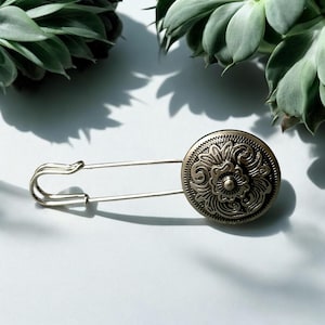 Könnte beinhalten: Eine silberfarbene Sicherheitsnadel mit einem dekorativen, kreisförmigen Knopf im antiken Stil. Der Knopf hat ein geprägtes Blumenmuster. Die Nadel liegt auf einer weißen Oberfläche, mit grünen Sukkulenten im Hintergrund.