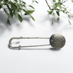 Könnte beinhalten: Sicherheitsnadel in Silberoptik mit einem dekorativen, kreisförmigen Kopf mit einem komplizierten, sich wiederholenden Muster. Die Nadel befindet sich vor einem weißen Hintergrund, mit einem Zweig grüner Blätter im oberen Teil des Bildes.