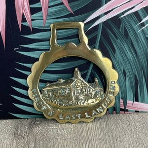 Könnte beinhalten: Ein Messing-Pferdegeschirr mit gewelltem Rand und einer rechteckigen oberen Schlaufe. In der Mitte befindet sich ein detailliertes Relief eines Gebäudes mit den Worten "FIRST & LAST LAND" am Rand.