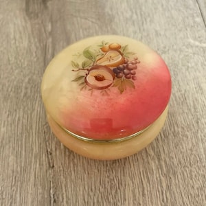 May include: A round, peach-colored trinket box with a lid featuring a painted design of fruits, including peaches, grapes, and pears. The box has a gold-colored rim and sits on a wooden surface.
