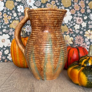 May include: A ceramic pitcher with a ribbed texture and a brown glaze featuring vertical streaks of muted green. The pitcher has a curved handle and a wide, flared rim. It is positioned alongside two decorative pumpkins on a wooden surface.