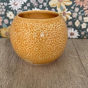 May include: A spherical ceramic bowl with a textured, speckled orange and yellow glaze. The bowl has a smooth, rounded rim and a glossy finish that catches the light, creating a subtle variation in the depth of the orange color across the surface.
