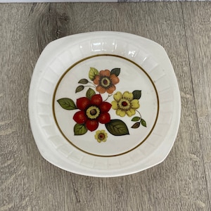 May include: A white ceramic plate with a floral design. The flowers are red, yellow, and orange with green leaves. The plate has a raised rim and a scalloped edge.