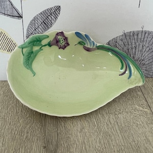 May include: A pale green ceramic dish with an unusual, flowing form. It features a floral design with purple flowers and green leaves. The rim is decorated with blue and green accents. A decorative piece.