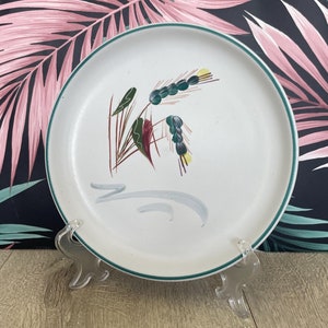 Könnte beinhalten: Weißer Keramikteller mit grünem Rand und floralem Design. Der Teller zeigt eine zentrale Illustration von stilisierten Weizenähren und Blättern in Grün-, Braun- und Burgundertönen. Der Teller wird auf einem transparenten Kunststoffständer präsentiert.