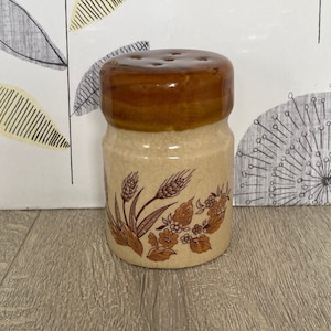 May include: A ceramic salt shaker with a brown lid and a beige body. The body is decorated with a brown wheat and floral pattern. The shaker has a classic, vintage design, perfect for kitchen decor.