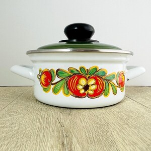 May include: A white enamel cooking pot with two side handles and a matching lid featuring a black knob. The exterior of the pot is decorated with a hand-painted floral motif in red, yellow, and green colors, encircling the center of the vessel.