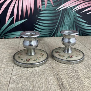 May include: Two vintage candle holders. Each has a silver-colored cup and stem, atop a round base with a cream-colored background and a floral pattern in red, pink, and green. The bases are trimmed with silver metal.