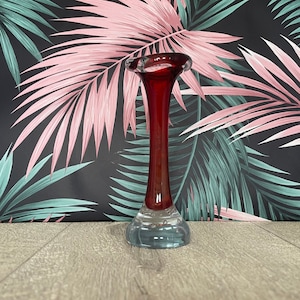 May include: A slender glass vase with a flared top and a long, elegant body. The vase has a clear, rounded base that leads up to a deep red colour. The background features a tropical leaf pattern in pink and teal.