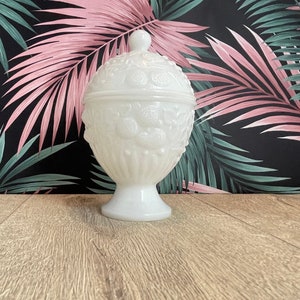 Peut inclure: Un pot à bonbons blanc avec un couvercle et un pied. Le pot présente un motif gaufré texturé de fleurs et de feuilles. Le fond présente un fond noir avec des feuilles tropicales roses et vertes.