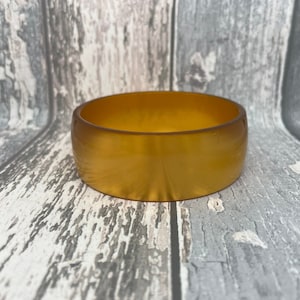 May include: A wide, amber-colored bangle bracelet. The bracelet has a smooth, polished surface and a rounded shape. The color is a warm, golden hue, and the bracelet appears to be made of a solid material. This accessory is suitable for everyday wear.