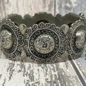 May include: Silver-coloured metal cuff bracelet with intricate floral and scrollwork designs. The bracelet features three large, circular medallions with embossed patterns and a delicate chain. The cuff is set against a weathered wood background.
