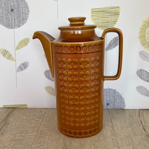 Könnte beinhalten: Eine hohe, braune Keramik-Kaffeekanne mit Deckel und Griff. Die Kanne hat ein dekoratives Muster aus orangefarbenen Blumen und Kreisen auf der Vorderseite. Der Hintergrund ist eine helle Wand mit einem floralen Design. Die Kanne ist etwa 25 cm hoch.
