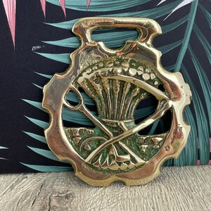 May include: A gold-colored brass horse brass with a green patina. The design features a thistle and crossed golf clubs.
