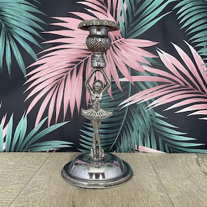 May include: Silver-colored candlestick holder featuring a ballerina figurine. The candlestick has a round base and a decorative top. The ballerina is holding the top of the candlestick. The background is a black and pink floral pattern.