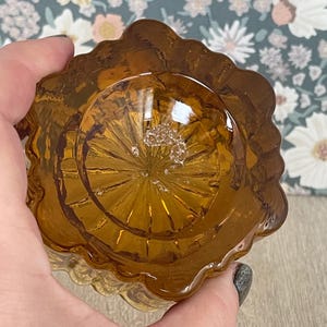 Könnte beinhalten: Bernsteinfarbene Glasschale mit gewelltem Rand. Die Schale hat ein strukturiertes Inneres und wird von einer Hand gehalten. Der Hintergrund zeigt ein Blumenmuster. Die Schale ist wahrscheinlich Vintage oder antik.