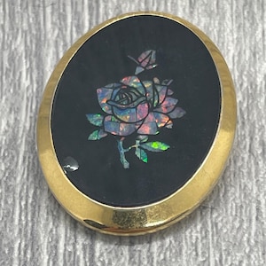 May include: Oval gold-tone brooch with a black background. The brooch features a rose design made of iridescent, colourful material. The rose has pink, green, and blue hues. A vintage accessory.