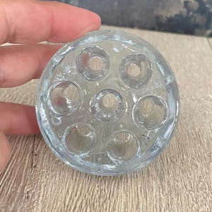 May include: A clear glass flower frog with multiple circular holes. The frog is round and appears to be made of thick glass. The holes are evenly spaced and designed to hold flower stems in place.