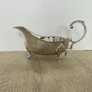 May include: A silver gravy boat with a curved spout and a decorative handle. The boat has three small feet and shell-shaped accents on the sides. The gravy boat is made of a shiny, reflective metal.