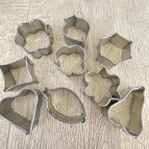 Peut inclure: Un ensemble de neuf emporte-pièces en métal de différentes formes, dont un cœur, une feuille et une fleur. Les emporte-pièces sont en métal argenté et sont disposés sur une surface en bois brun clair. Les formes sont variées, pour la pâtisserie.