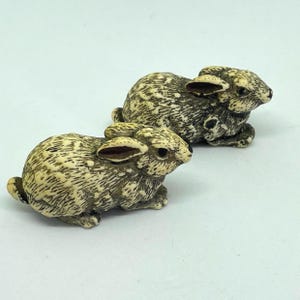 May include: Two miniature rabbit figurines. The rabbits are depicted in a seated pose, with their ears erect. They feature a mottled cream and brown pattern, with dark eyes and ears. The figurines are likely crafted from a hard material.