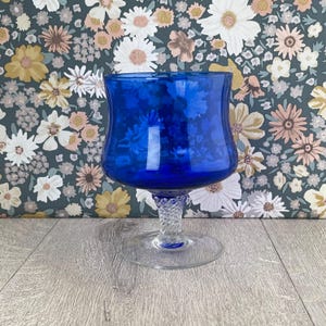 May include: A vibrant blue glass goblet with a flared bowl and a clear, twisted stem. The goblet sits on a clear, circular base. The background features a floral pattern with white, yellow, and peach flowers on a dark green background.