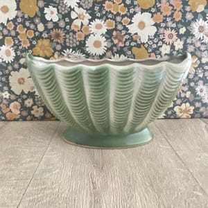 May include: A ceramic planter with a scalloped, wavy rim and a ribbed, vertical texture. The exterior features a gradient of sage green and off-white tones, while the base is a solid, smooth oval shape that supports the wide, bowl-like vessel.