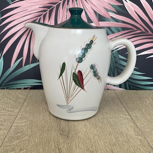 May include: Cream ceramic pitcher with a green lid and handle. The pitcher is decorated with a floral design in green, red, and brown. The background features a pink and green tropical leaf pattern.