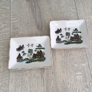 以下が含まれることがあります： 金色の縁取りが付いた、正方形の白い陶器の小皿が2つ。それぞれのお皿には、建物、木、鳥が描かれたカラフルな風景画が描かれています。小皿は、明るい灰色の木製の表面に置かれています。