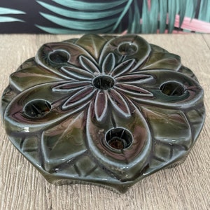 以下が含まれることがあります： 暗い緑色のセラミックキャンドルホルダー、花の模様。キャンドルホルダーは円形で、キャンドル用の複数の開口部があります。デザインは、花びらのような形に囲まれた中心の花を特徴としています。
