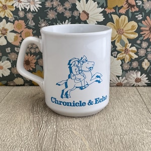 May include: A white ceramic mug featuring a blue graphic of a person riding a horse and the text Chronicle & Echo printed in blue. The mug has a curved handle on the left side and a smooth, cylindrical body with a wide rim and a flat base.