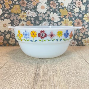 May include: A white, round bowl with two handles. The bowl is decorated with a colourful floral pattern around the rim, featuring yellow, blue, orange, and red flowers. The bowl is set against a floral patterned background.