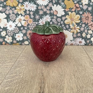 May include: A ceramic strawberry-shaped jar with a green leaf-shaped lid. The jar is a vibrant red with small white seed details. The lid has a green stem. The jar is likely used for storing small items or as a decorative piece.