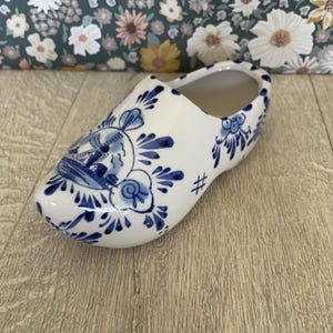 May include: A white ceramic clog featuring blue hand-painted floral patterns and a detailed illustration of a windmill on the toe. The shoe-shaped decorative object has a glossy finish and a hollow interior, showcasing traditional blue and white motifs.