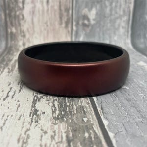 May include: A wide, round, maroon bangle bracelet with a black interior. The bracelet is made of a smooth, glossy material.