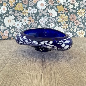 May include: A dark blue glass bowl with white speckles. The bowl has a unique, asymmetrical shape and sits on a small, round base. The interior of the bowl is a deep, vibrant blue. The background features a floral pattern.