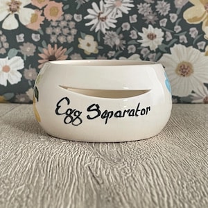 May include: A cream-coloured ceramic egg separator with a smooth, rounded body and a horizontal slit opening. The words Egg Separator are written in black cursive script on the front surface of the bowl, which features a glossy finish.