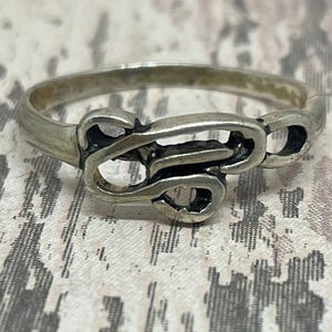 May include: A silver-toned ring with an openwork design. The ring features a series of interconnected, stylized shapes, creating an intricate pattern. The metal has a slightly aged appearance, adding to its vintage appeal. The ring is a piece of jewelry.