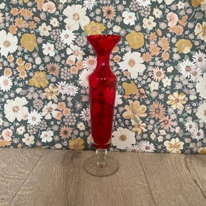 May include: A tall, vibrant red glass vase featuring a slender neck that flares into a wide, circular rim. The vase is supported by a clear glass stem and a circular base, showcasing a smooth, polished finish throughout the entire structure.
