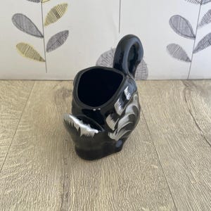 May include: A black ceramic swan-shaped planter with a silver painted design. The planter has a curved neck and a hollow interior for holding plants or other decorative items. The swan's body is detailed with silver brushstrokes.