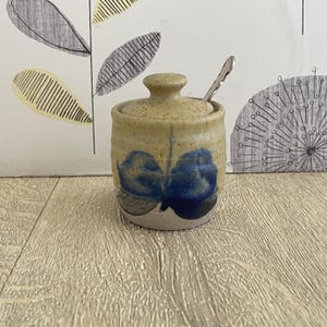 May include: A small ceramic sugar bowl with a lid and a spoon. The cylindrical bowl has a tan base and blue floral designs. The lid is tan with a small knob. A silver spoon rests inside the bowl.