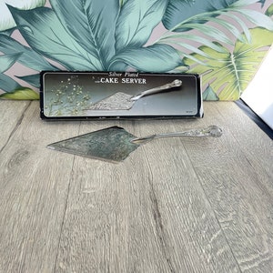 Könnte beinhalten: Versilberter Tortenheber mit dekorativem Blumendesign. Der Heber wird mit seiner Originalverpackung präsentiert, auf der "Silver Plated CAKE SERVER" steht. Der Hintergrund zeigt ein tropisches Blättermuster.