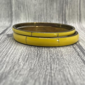 May include: Two circular bangles featuring a bright yellow enamel finish over a metallic base. Each bangle is divided into segments by thin, dark vertical lines that create a repeating pattern around the circumference of the jewellery pieces.