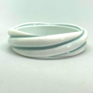 May include: A pale blue and white ceramic bangle bracelet. The bracelet features a twisted design with a glossy finish. The bracelet is a piece of jewellery.