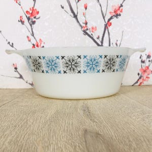 May include: White Pyrex casserole dish with a decorative band of blue and black snowflake patterns. The dish has two handles. A vintage kitchenware item. The background features a floral pattern.