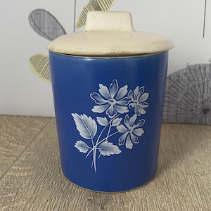 May include: A blue ceramic canister with a cream-colored lid. The canister features a white floral design. The lid has a rectangular handle. The canister is likely used for storing dry goods.