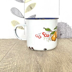 May include: White enamel mug with a blue rim and a floral design featuring pears, cherries, and flowers.
